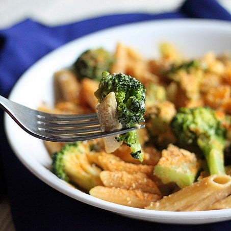 A white bowl of Penne and Broccoli in Doritos cream sauce on a blue cloth. A fork in the foreground with some penne and broccoli on it.