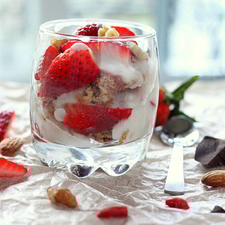 A glass of Spiced Oat Almond Gluten-free Cake, in Strawberry Nuts Goji berry parfait on a fabric background with pieces of strawberry scattered around