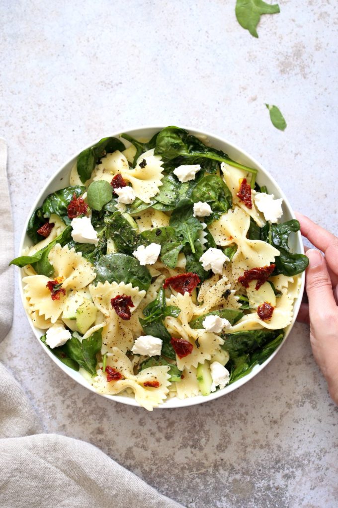 A bowl of Tuscan Pasta Salad on a light worktop. A hand holds the side of the bowl