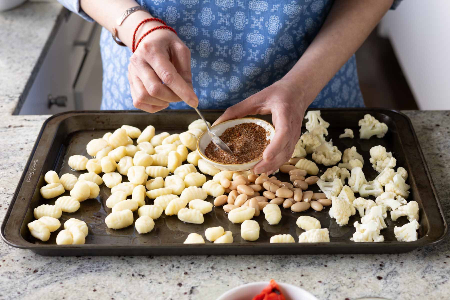 gnocchi, beans, and cauliflower on the baking sheet while a hand mixes the spices in a bowl